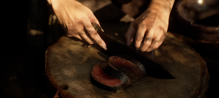 Close-up of hands slicing fresh fish on a wooden cutting board with a sharp knife. The fish has silvery skin and pinkish flesh, glistening under warm kitchen light.