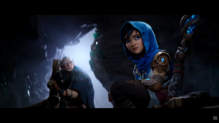 Kena sits in a dimly lit cave, wearing a blue hooded cloak and holding her staff. Her expression is serious as she looks off-screen. Behind her, an older man with a beard and armor stands guard, holding a weapon.
