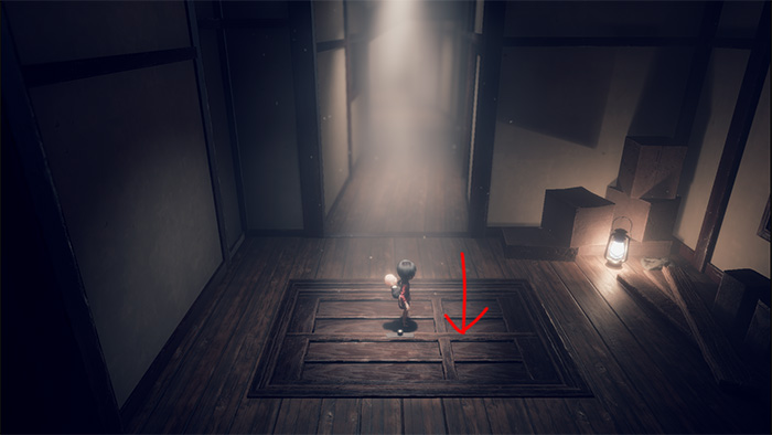 The player character stands on a wooden floor panel with a red arrow pointing downward, indicating an interactive area. A lantern glows faintly to the right, casting shadows in the dim room. The environment suggests a hidden mechanism or puzzle element beneath the floor.