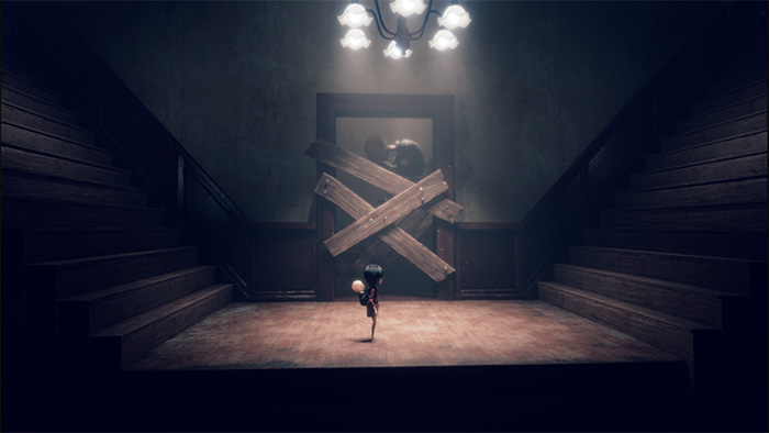A young girl character stands at the bottom of a dimly lit staircase, facing a door blocked by two wooden planks arranged in an "X" shape. Above, a chandelier casts soft light, creating shadows on the worn walls. The scene suggests a locked or forbidden passage.