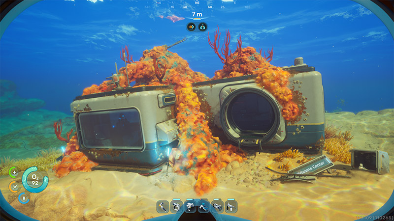 An abandoned modular structure rests on the seafloor, heavily encrusted with vibrant orange coral and feathery anemones; a small sign reading “Welcome Center” lies half-buried nearby.