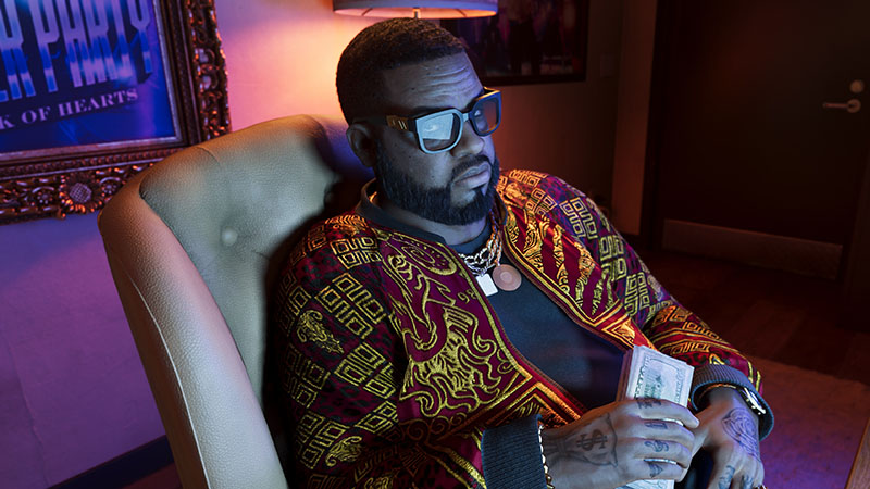 GTA6 Boobie Ike reclining in a cream leather armchair—wearing oversized black-framed glasses, a richly patterned maroon-and-gold embroidered jacket over a dark shirt, layered gold chains, and a pendant. He holds a stack of cash; visible tattoos on both hands.
