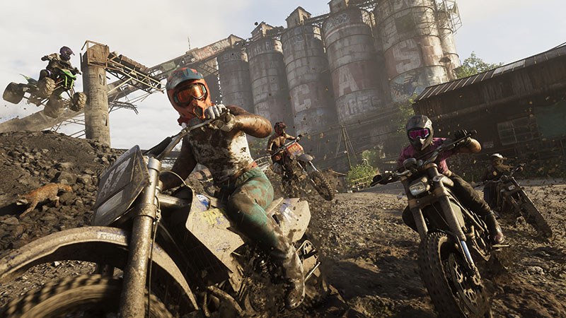 GTA6 official scene: Four dirt bikers racing through a muddy industrial ruin—foreground rider in orange goggles and mud-splattered gear leans sharply on a white off-road bike; others trail behind amid flying debris, with rusted silos and conveyor structures in the background.
