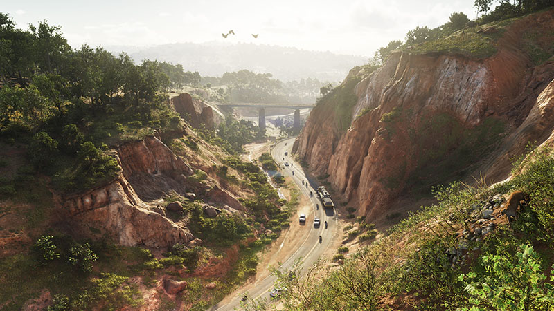 GTA6 official scene: A winding two-lane highway cuts through a sun-drenched canyon with eroded red sandstone cliffs, lush green vegetation on slopes, distant bridge and city skyline under hazy daylight, and multiple vehicles in motion — including cars, motorcycles, and a bus — with birds flying overhead.