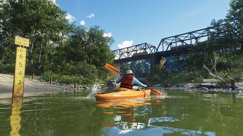 GTA6 official scene: A kayaker in red life vest paddling an orange Crest kayak through shallow green river water, with a detailed flood gauge sign (marked in feet) reflecting on the surface, a rusted steel truss bridge overhead carrying a freight train, and lush riparian vegetation.