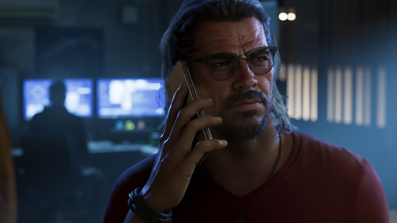 GTA6 protagonist Raul Bautista on a phone call in a dimly lit control room: a middle-aged man of Latinx descent with long dark hair tied back, a full beard, and rectangular metal-framed glasses. He wears a maroon V-neck shirt, a dark beaded bracelet, and a pendant necklace.