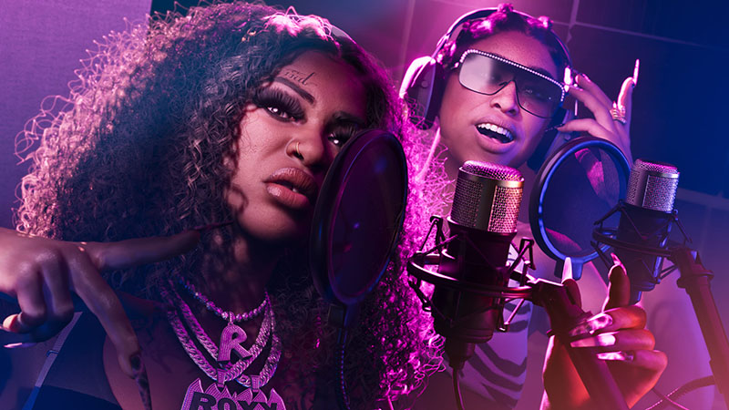 GTA6’s fictional rap duo Real Dimez in a neon-lit studio: two Black women singing into professional mics with pop filters. Left: voluminous curly afro, bold makeup, layered gold chains including an “R” pendant, forehead tattoo reading “real”. Right: oversized reflective sunglasses, striped top, hand near mic, expressive pose.