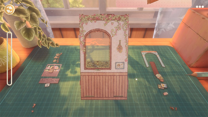 On a green grid mat, an arched paper book nook with a wooden base is being assembled, surrounded by a few loose pieces; a potted plant on the left, a box and pencil on the right, with a sunlit window in the background.