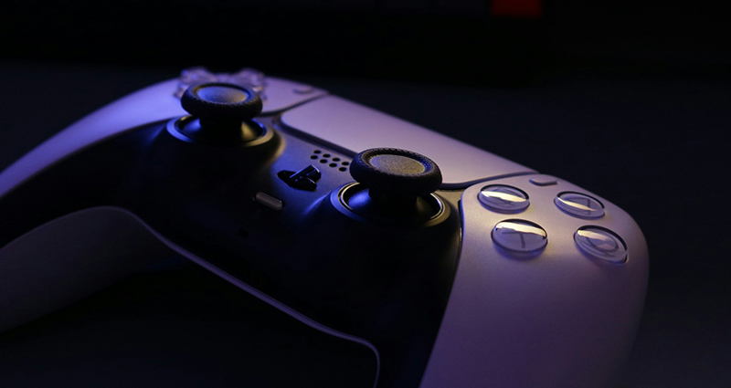 Close-up studio photo of a white PlayStation 5 DualSense controller, lit with cool blue-purple ambient light, highlighting the analog sticks, touchpad, and action buttons.