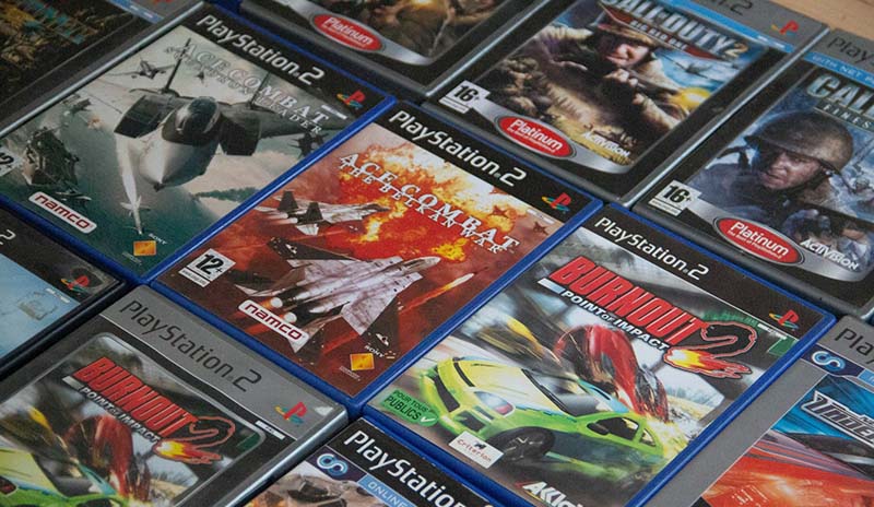 Close-up photo of stacked PlayStation 2 game cases, including titles like "Ace Combat" and "Burnout", some labeled "Platinum"; shows physical disc-based gaming era.